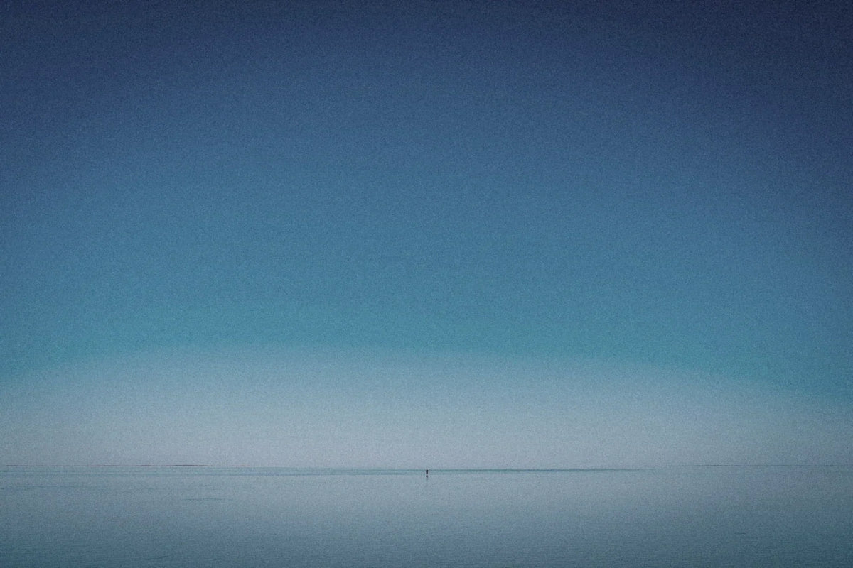 A lone figure standing in shallow water against a wide blue horizon.