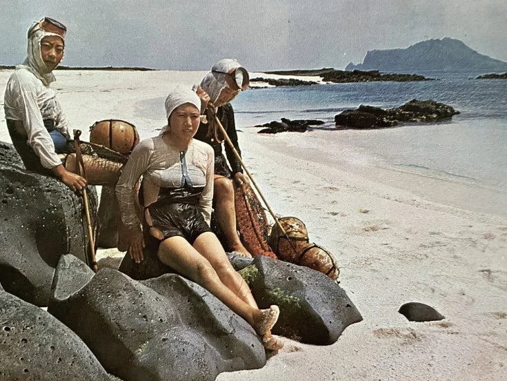 Haenyeo divers resting on volcanic rocks at Jeju in 1971, floats and hooks beside them.