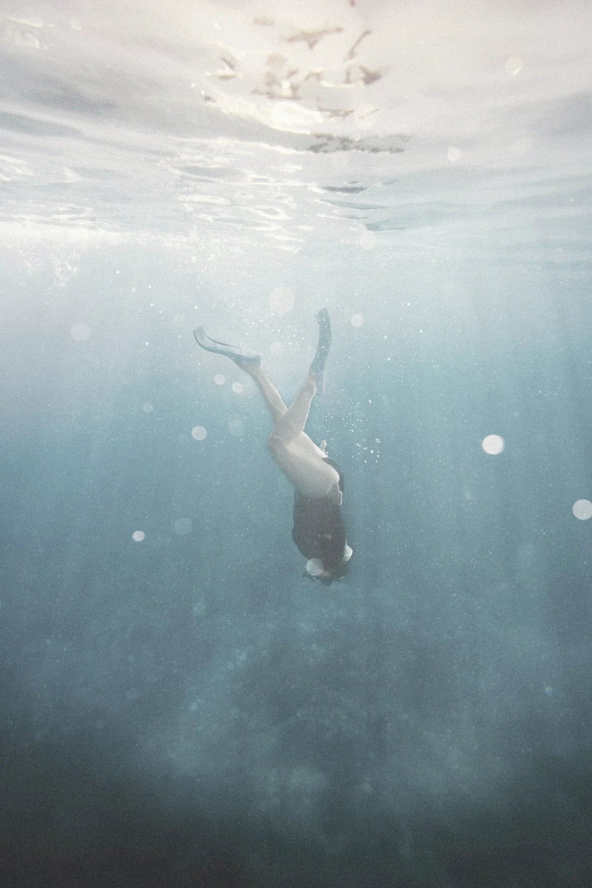 A swimmer moves down through open salt water, seen from below the surface.