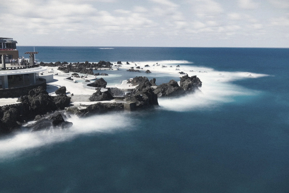 Rock pools walled off from the Atlantic by black volcanic rock, white water breaking around them, Portugal.