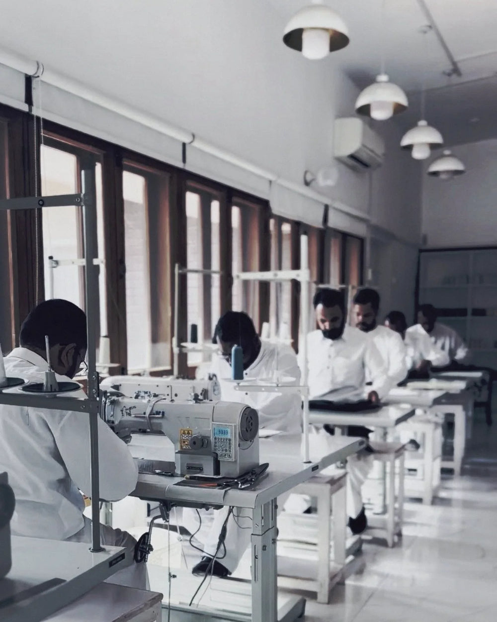 Craftsmen at their machines along the atelier floor, garden light through the windows