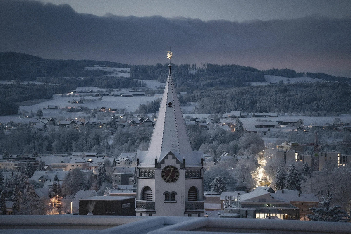 An alpine village under snow at dusk, a church spire and lit windows, mountains behind.