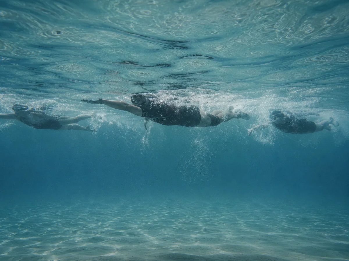 Swimrun athletes swimming in a line through clear turquoise water, seen from below the surface.