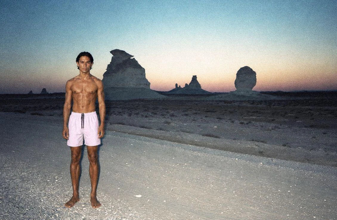 A man in pale swim shorts standing barefoot in Egypt's White Desert at dusk, chalk formations behind him.