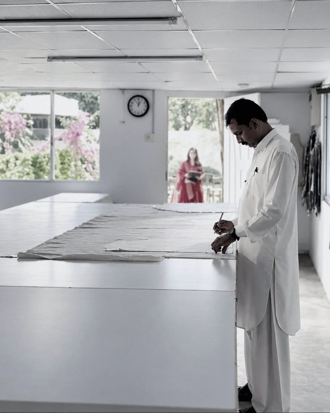 The cutting room: a cutter marks cloth at the table, a colleague at the garden door